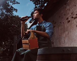 Indian brewer in uniform sitting on a wooden barrel and drink craft beer.
