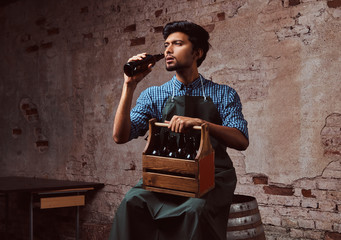Indian brewer in uniform sitting on a wooden barrel and drink craft beer.