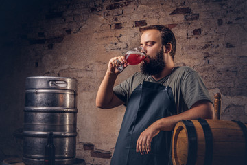 Brewer in apron standing near barrels and drinks craft beer at old brewery factory.
