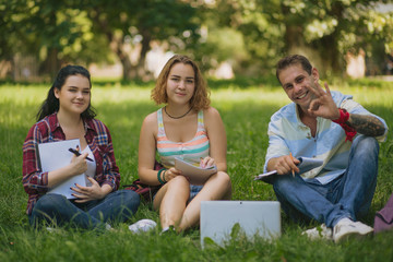 Naklejka premium Students sit on the grass and doing their homework. Guy with a tatto shows the ok gesture. Two girl hold the notebooks. Laptop is on the grass.