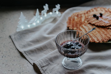 The concept of home winter comfort. Delicious crispy homemade waffles with jam in a glass bowl on the table at the winter window. In the background lights Christmas garland.