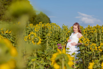 Fototapeta premium A young pregnant girl in a white dress in sunflowers field. Happy moments of pregnancy. Authentic lifestyle image. place for text