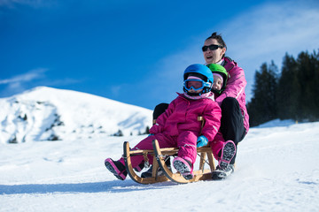 Mother and children having fun on sledge with mountain scenery in background. Active mom and kids with safety helmets. Winter sport for family. Little kids outside, swiss Alps, healthy lifestyle  - obrazy, fototapety, plakaty