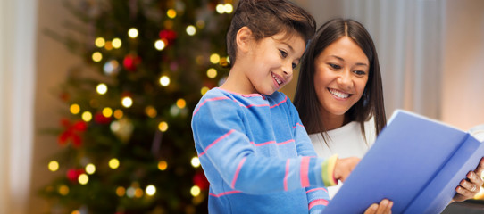 family, holidays and people concept - happy mother and daughter reading book over christmas thee lights background