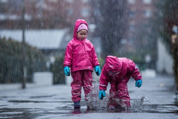 Girls are having fun in water on street in cold autumn day, girls splashing water in rain, happy and cheerful girls enjoying cold weather, kids in pink rain coats and rubber boots 