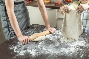 Two adult girl and the child cook dough. Dough is spread out on the big table sprinkled with flour. The child has lifted the rolled dough over a table, and the girl rolls dough with a rolling pin