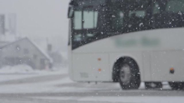 SLOW MOTION, DOF: White bus slows down at a bus stop during a snowy winter day. Bad weather gathers over the city streets and covers them in snow. Tourist coach stops due to bad weather conditions.
