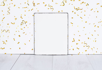 White empty board standing on grey wooden floor against white isolated wall with golden party confetti mock up for birthday