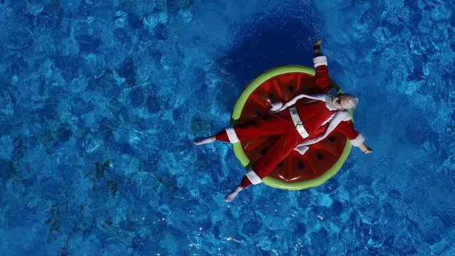 Aerial view: Santa lie on the watermelon inflatable ring and calling to come makes a hand gesture. Concept of satisfaction and xmas vacation. Cheerful man celebrate life, summertime and happiness