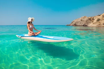 Foto op Canvas Young beautiful woman meditating in a sea at SUP paddleboarding. Healthy lifestyle. Girl in yoga pose relaxing in calm water. © kuznetsov_konsta