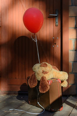 Pluszowy piesek leżący na walizce z czerwonym balonem przed drzwiami domu - obrazy, fototapety, plakaty