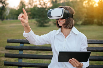 Fototapeta premium Young businesswoman using virtual reality goggles while sitting on a bench in a city park and working.