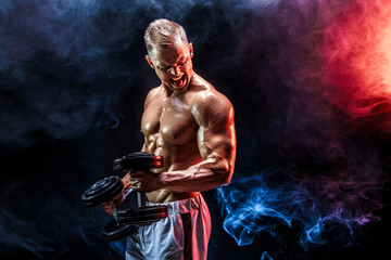 Fototapeta na wymiar Topless man exercising biceps with dumbbells posing in studio fuul of colored smoke