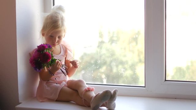 The young ballerina sits on the window and holds flowers in her hands