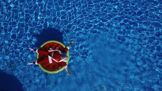 Aerial view: Santa lie on the watermelon inflatable ring and calling to come makes a hand gesture. Concept of satisfaction and xmas vacation. Cheerful man celebrate life, summertime and happiness