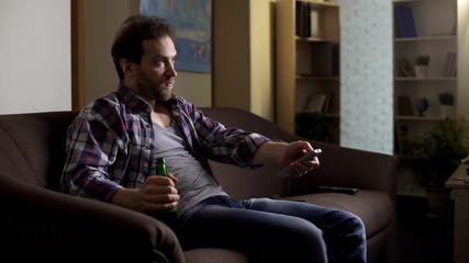 Guy sitting on sofa with beer bottle in hand, using remote control to switch TV