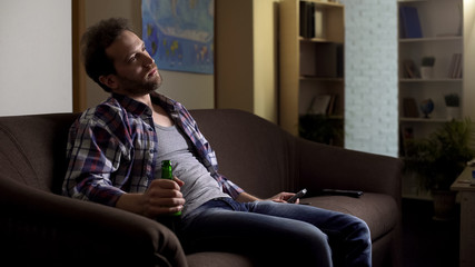 Fototapeta premium Grubby man sitting on sofa with beer bottle in hand and watching TV, alcoholism