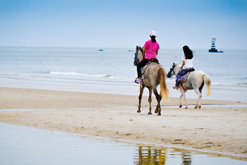 Back of young asian girls ridding horse on the beach with lighthouse in far background in the sea.