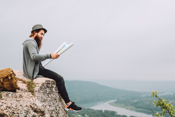 Young bearded tourist sits on a high rock.The guy holds the map.Behind the guy green trees, forest and a long river.