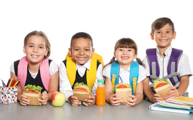 Schoolchildren with healthy food and backpacks sitting at table on white background