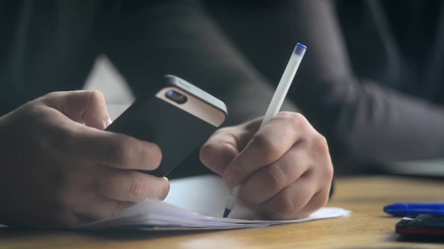 Close-up a Man making Notes with a Pen, Paper and Mobile Smart Phone in Hands