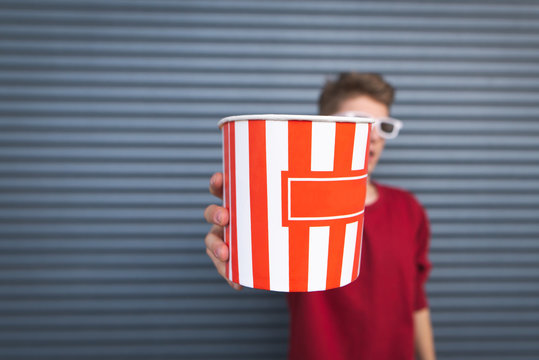 Guy stands on a dark background, holds a bowl of popcorn in his hands and shows him in the camera. Cup with popcorn in the hands of a man close-up.