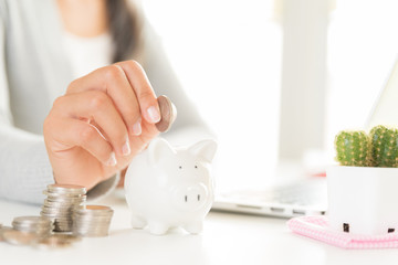 Woman hand putting money coin into piggy bank with stack of coins. Computer calculator on office table. Saving money wealth and financial concept.