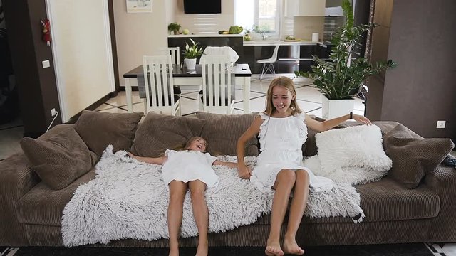 Attractive two girls fun falling and lies on the sofa spreading their arms in the living room at their home. Beautiful, happy girls in white dresses standing on the floor and have fun falling back on