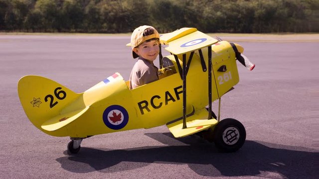 Cinemagraph of a boy in a toy biplane on the tarmac with forest in the background.