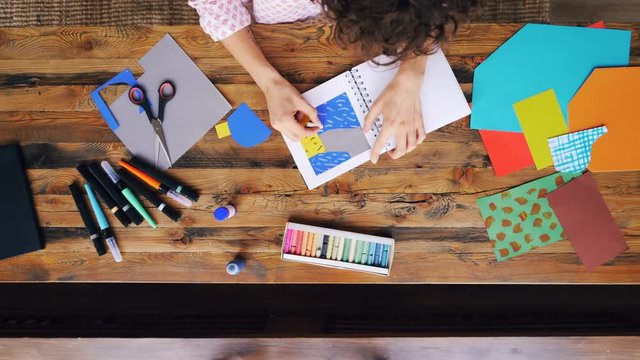 Zoom-out of creative brunette making paper collage drawing with crayons sitting at desk in home office. Artists, professionals and workplace concept.
