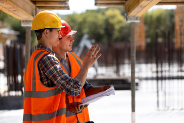 Two civil engineers dressed in orange work vests and helmets discuss the construction process on the building site near the wooden constructions and steel frames