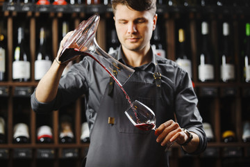 Bartender pours red wine in glass from big transparent vessel