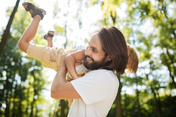 Dark-haired father with beard dressed in the white t-shirt is holding in the arms his blond son and smiling on a summer day in the park.