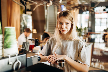 A pretty smiling slim girl,wearing casual clothes,holds some ground coffee and looks at the camera in a cozy coffee shop.