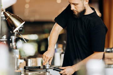 A stylish young man with beard,wearing casual clothes,cooks coffee in a coffee machine in a cozy coffee shop.