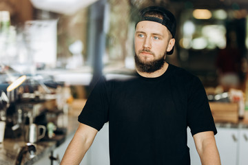 A young stylish man with beard,wearing casual clothes,stands thoughtfully in a cozy coffee shop.