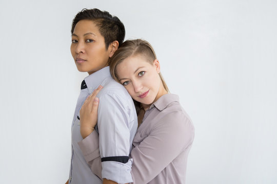 Two Serious Women Hugging And Looking At Camera. Multiethnic Homosexual Couple Posing. Lesbian Couple Concept. Isolated Side View On White Background.