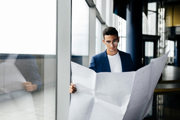 Fototapeta premium Architect in stylish clothes holds sheet with drawing in his hand and talks by phone on the background of a modern glass multistory building