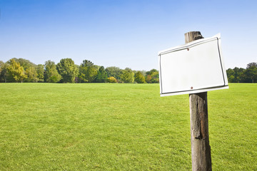 Wooden blank sign indicating against a green mowed lawn with trees and sky on background in the countryside - image with copy space