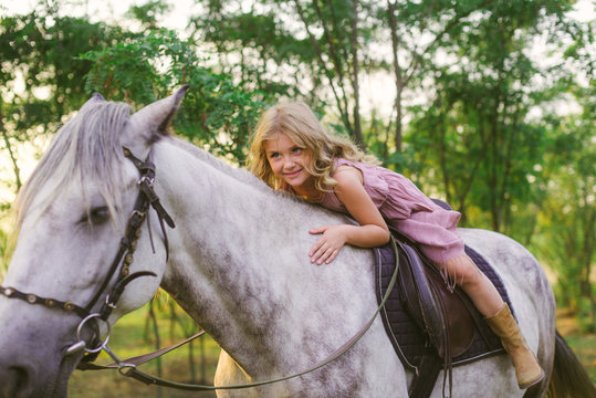 Little cute girl with light curly hair in a straw hat riding a horse at sunset on a sunny warm autumn day
