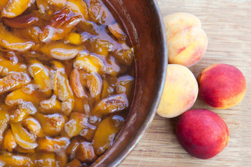Fresh bright peaches and nectarins lay on the kitchen desk close to the copper jar, full of peach jam. Close up, outdoor.