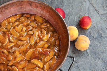 Fresh bright peaches and nectarins lay on the stone bricks, close to the copper jar, full of peach jam. Close up, outdoor.