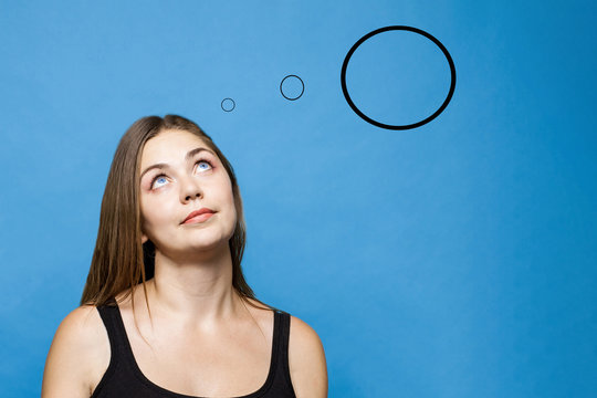 Young causacian woman with blue eyes up thinks or dreams about something. Three rounds, each more than previous one, fly from her head, all are empty, dark hair, blue background, studio. Copy space.
