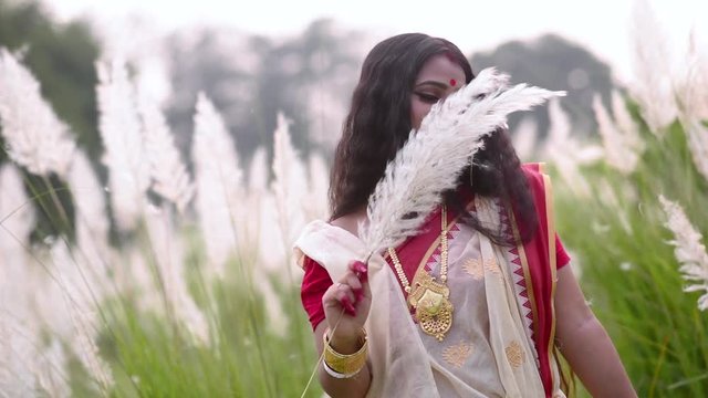 A cute and carefree newly wed Indian woman is happy and plays with white flowered grass in a field on a windy day at sunset or surise