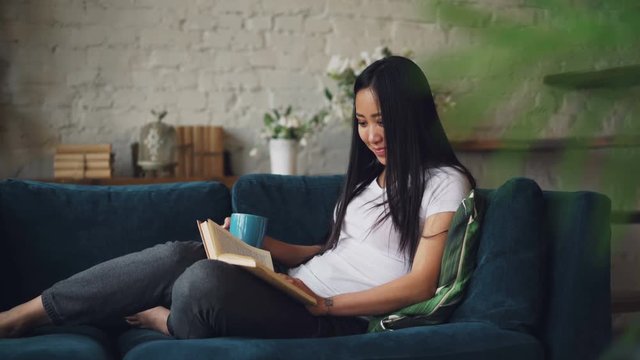 Beautiful Asian woman is reading book enjoying funny story and laughing sitting on sofa in cozy loft style apartment and resting. Nice furniture and plants are visible.