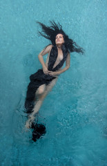 schöne Frau mit dunklen Locken in schwarzem Kleid, Abendkleid schwebt schwerelos elegant glücklich schwimmend in türkis blauem Wasser im Pool
