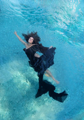 schöne junge Frau in schwarzem Kleid, Abendkleid schwebt schwerelos elegant schwimmend im Wasser im Pool dunkel braune lockige Haare treiben fächerförmig  in türkisem Wasser