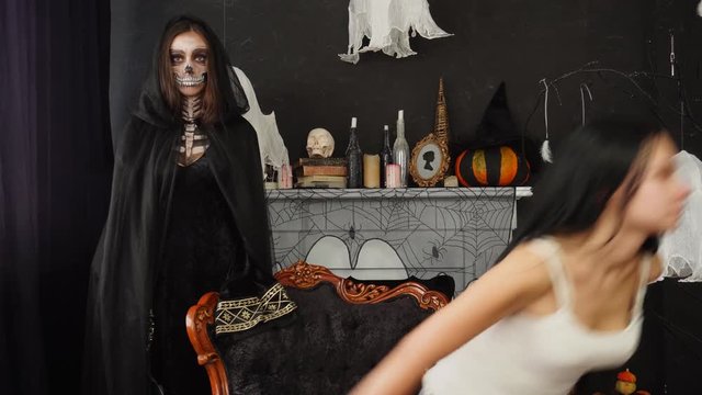 Young attractive brunette sitting in retro chair afraid of Death Reaper standing behind spider web decorated fireplace