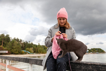 Close-up of woman hands knitting  pink  wool hat with needle,    next is a beautiful gray cat against the background  of the sea  in sunny autumn day . Freelance creative working and living concept