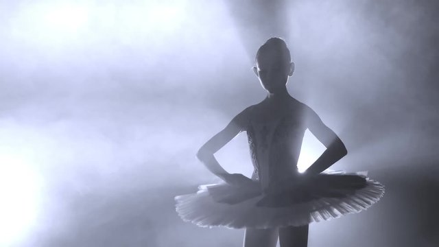 Ballerina's silhouette turning around. ballerina with hair in a bun is dancing on a scene, against bright light of soffit and smokescreen